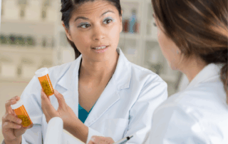 A female pharmacy technician explains prescription bottles to a colleague while reviewing notes in a pharmacy environment.