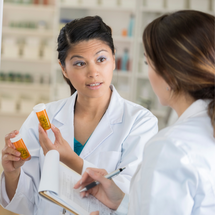 A female pharmacy technician explains prescription bottles to a colleague while reviewing notes in a pharmacy environment. A female pharmacy technician explains prescription bottles to a colleague while reviewing notes in a pharmacy environment.