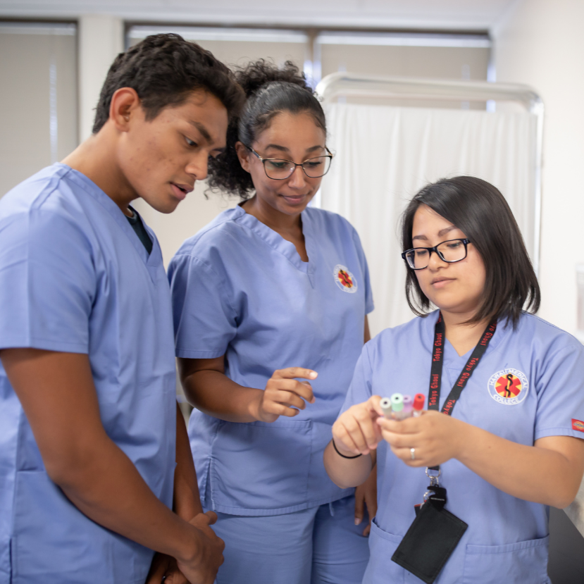 Three medical assistant students in blue scrubs participate in hands-on training, examining blood collection tubes together in a clinical classroom setting. Three medical assistant students in blue scrubs participate in hands-on training, examining blood collection tubes together in a clinical classroom setting.